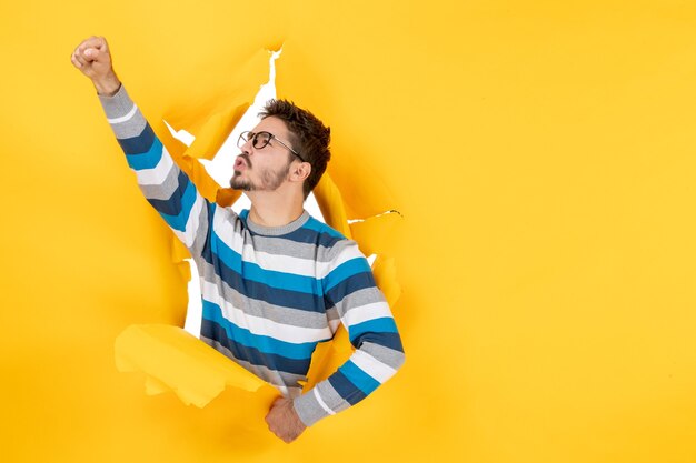 Vista frontal joven levantando su mano asomando a través del orificio en la pared de papel amarillo