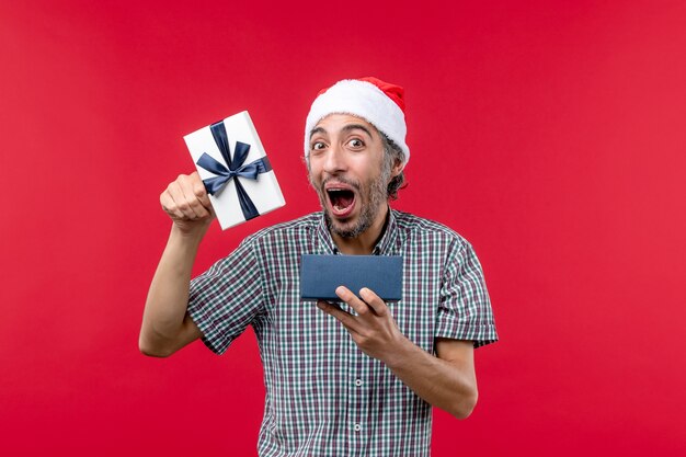 Vista frontal del joven macho abriendo regalo de Navidad en rojo claro