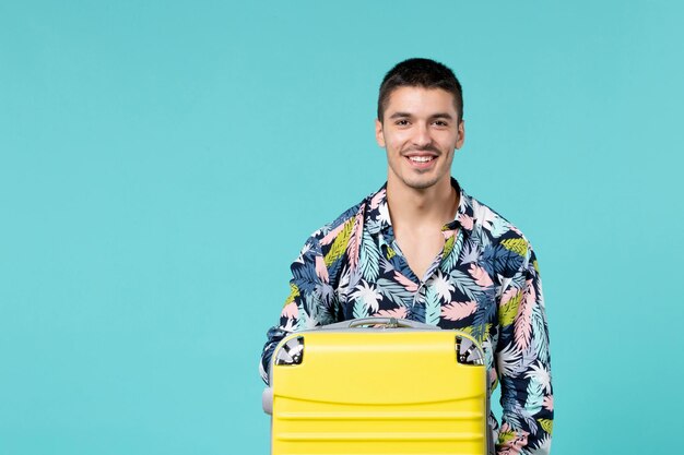 Vista frontal del joven macho con bolsa amarilla sosteniendo su sombrero y preparándose para el viaje en la pared azul