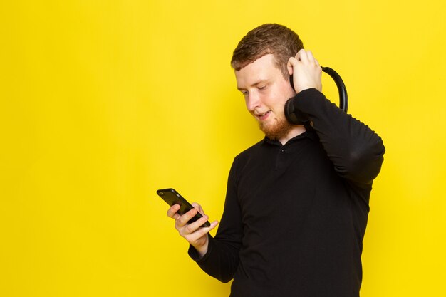 Vista frontal del joven macho en camisa negra escuchando música a través de auriculares con una sonrisa