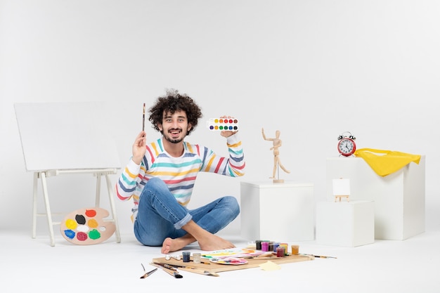 Vista frontal del joven macho dibujando imágenes con pinturas en la pared blanca