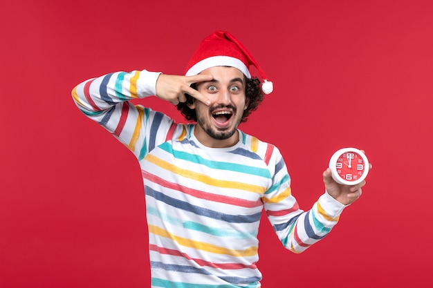 Vista frontal joven macho felizmente sosteniendo relojes en la pared roja tiempo de vacaciones de año nuevo macho rojo