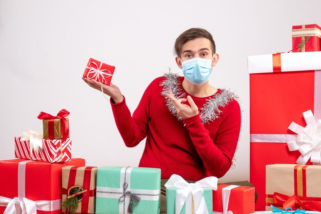 Vista frontal joven con máscara apuntando al regalo de Navidad sentado alrededor de regalos de Navidad