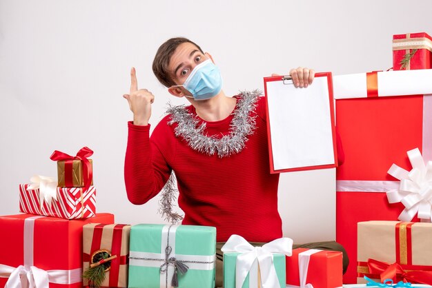 Vista frontal joven con máscara apuntando al techo sentado alrededor de regalos de Navidad