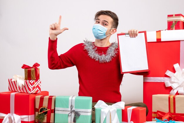 Vista frontal joven con máscara apuntando con el dedo hacia arriba sentado alrededor de regalos de navidad