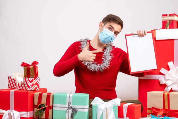 Vista frontal joven con máscara apuntando con el dedo portapapeles sentado en el piso blanco de regalos de navidad