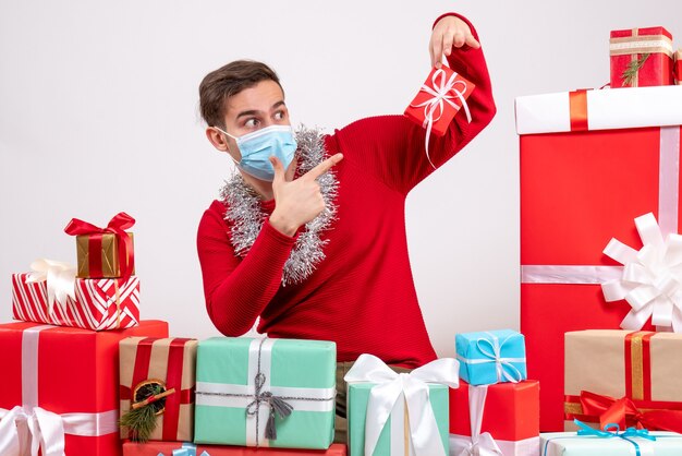 Vista frontal joven con máscara apuntando a regalo sentado alrededor de regalos de navidad