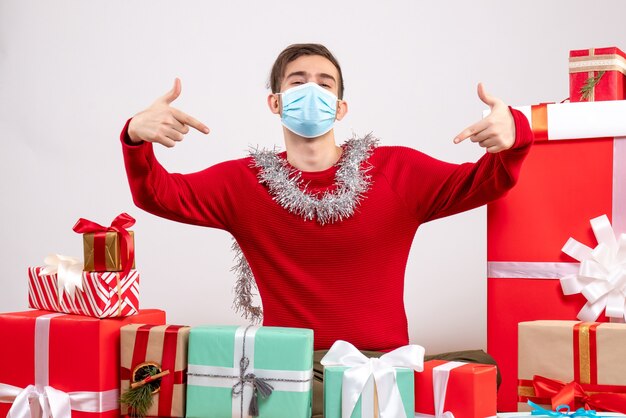 Vista frontal joven con máscara apuntando a sí mismo sentado alrededor de regalos de Navidad