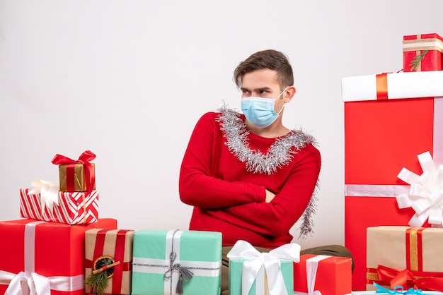 Vista frontal joven con máscara cruzando las manos sentado alrededor de regalos de Navidad