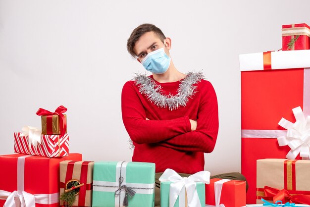 Vista frontal joven con máscara cruzando las manos sentados alrededor de regalos de Navidad