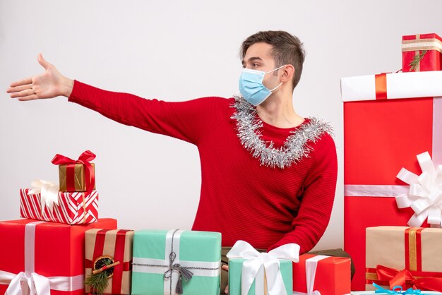 Vista frontal joven con máscara dando mano sentado alrededor de regalos de Navidad