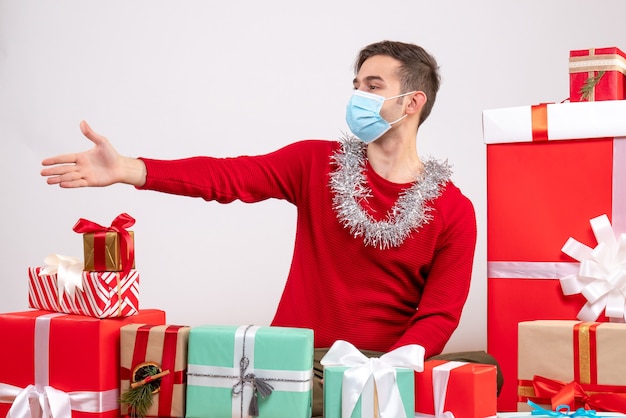 Vista frontal joven con máscara dando mano sentado alrededor de regalos de Navidad