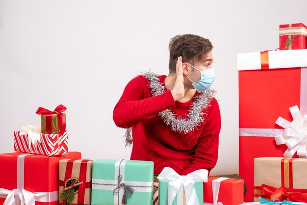 Vista frontal joven con máscara escuchando algo sentado alrededor de regalos de Navidad