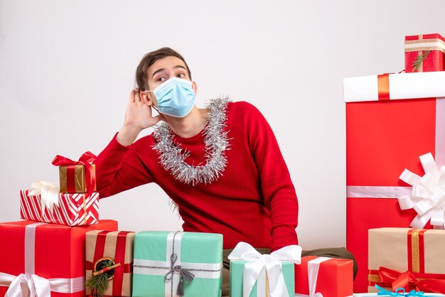 Vista frontal joven con máscara escuchando algo sentado alrededor de regalos de Navidad