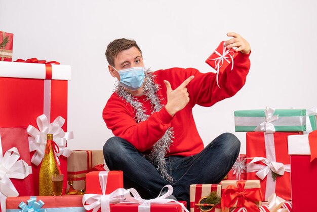 Vista frontal del joven en máscara estéril sentado alrededor de regalos de Navidad en la pared blanca