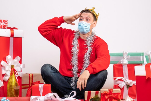 Vista frontal del joven en máscara estéril sentado alrededor de regalos de Navidad en la pared blanca