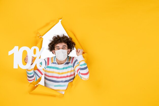 Vista frontal joven con máscara estéril sosteniendo la escritura en color amarillo venta de coronavirus covid de salud de foto