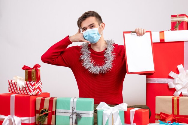 Vista frontal joven con máscara haciendo gesto de llamarme teléfono sentado en el piso regalos de navidad