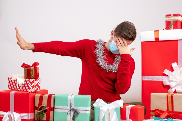 Vista frontal joven con máscara haciendo gesto de parada sentado alrededor de regalos de Navidad