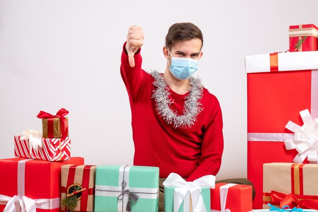 Vista frontal joven con máscara haciendo signo de pulgar hacia abajo sentado alrededor de regalos de Navidad