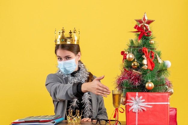 Vista frontal joven con máscara médica con corona dando mano árbol de Navidad y cóctel de regalos