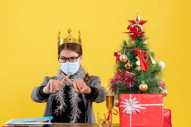 Vista frontal joven con máscara médica cruzando los dedos árbol de Navidad y cóctel de regalos