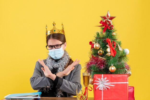 Vista frontal joven con máscara médica cruzando sus manos árbol de Navidad y cóctel de regalos