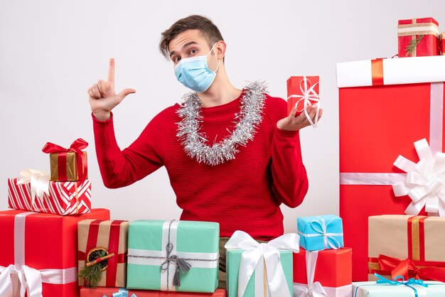 Vista frontal joven con máscara médica sentado alrededor de regalos de Navidad