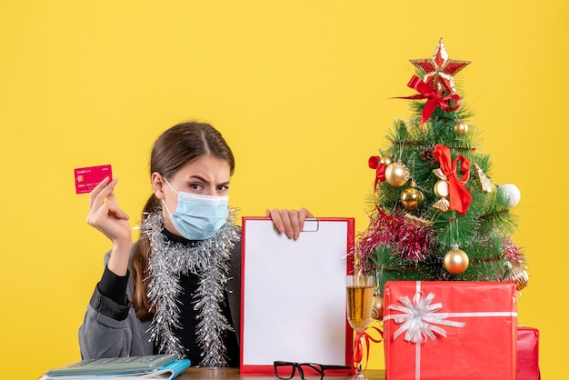 Vista frontal joven con máscara médica sentado en la mesa con tarjeta de crédito árbol de Navidad y cóctel de regalos