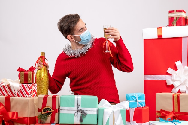 Vista frontal joven con máscara mirando champán sentado alrededor de regalos de Navidad