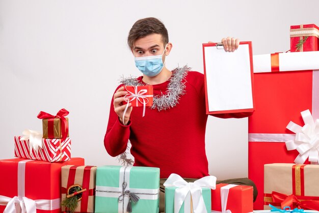 Vista frontal joven con máscara mirando regalo sentado alrededor de regalos de Navidad