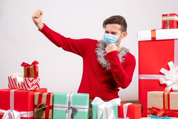 Vista frontal joven con máscara mostrando fuerza sentado alrededor de regalos de Navidad