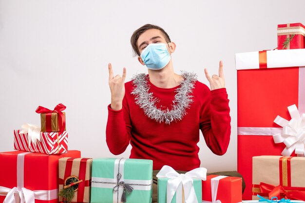 Vista frontal joven con máscara mostrando signo de rock sentado alrededor de regalos de Navidad