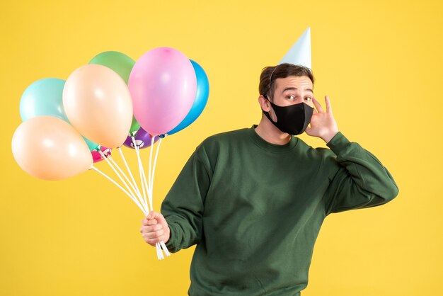 Vista frontal joven con máscara negra y globos de colores de pie sobre amarillo