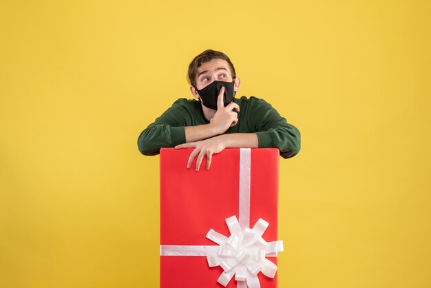 Vista frontal joven con máscara negra de pie detrás de caja de regalo grande en amarillo