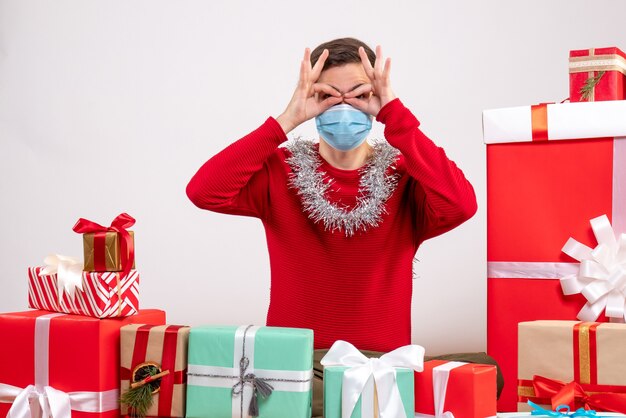 Vista frontal joven con máscara poniendo manos binoculares a los ojos sentado alrededor de regalos de Navidad