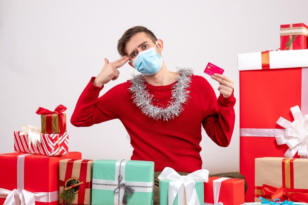 Vista frontal joven con máscara poniendo pistola de dedo en su sien sentado alrededor de regalos de navidad