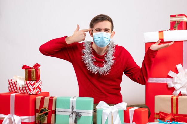 Vista frontal joven con máscara poniendo pistola de dedo en su sien sentado alrededor de regalos de navidad