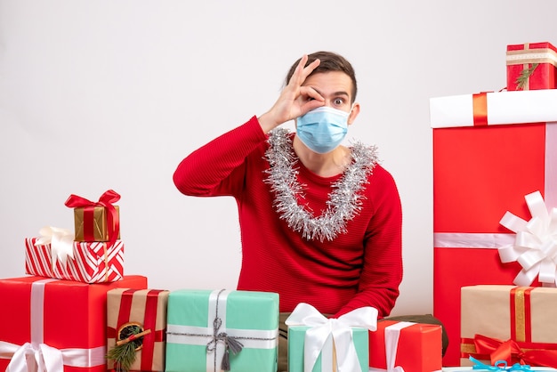 Vista frontal joven con máscara poniendo signo okey delante de su ojo sentado alrededor de regalos de Navidad