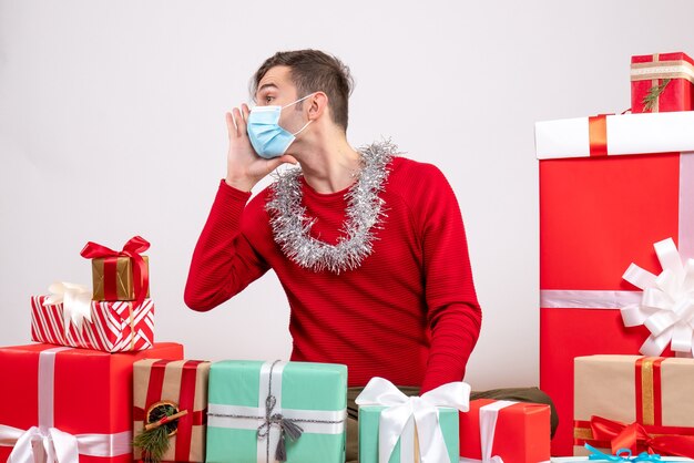 Vista frontal joven con máscara saludando a alguien sentado alrededor de regalos de Navidad