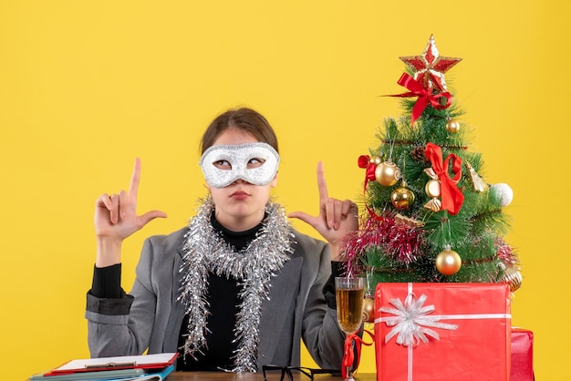 Vista frontal joven con máscara sentada en la mesa mostrando algo con los dedos árbol de Navidad y cóctel de regalos