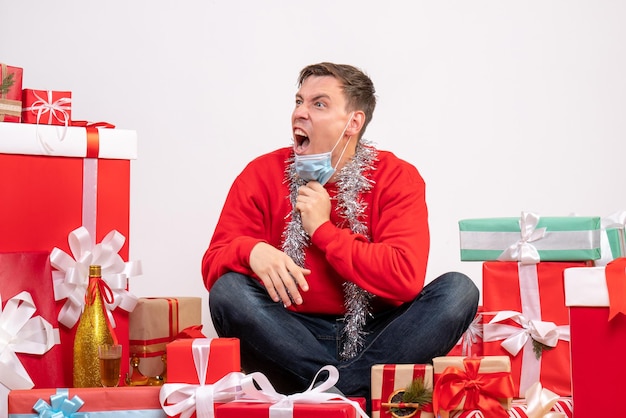 Vista frontal del joven con máscara sentado alrededor de regalos de Navidad gritando en la pared blanca