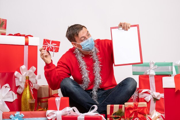 Vista frontal del joven con máscara sentado alrededor de regalos de Navidad con una nota en la pared blanca