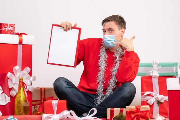 Vista frontal del joven con máscara sentado alrededor de regalos de Navidad con una nota en la pared blanca