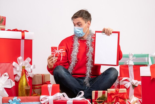 Vista frontal del joven con máscara sentado alrededor de regalos de Navidad con una nota en la pared blanca