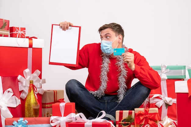 Vista frontal del joven con máscara sentado alrededor de regalos de Navidad con nota y tarjeta bancaria en la pared blanca