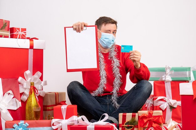 Vista frontal del joven con máscara sentado alrededor de regalos de Navidad con nota y tarjeta bancaria en la pared blanca