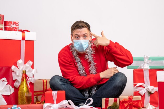 Vista frontal del joven en máscara sentado alrededor de regalos de Navidad en la pared blanca