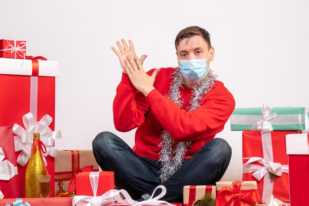 Vista frontal del joven en máscara sentado alrededor de regalos de Navidad en la pared blanca