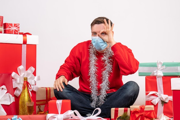 Vista frontal del joven en máscara sentado alrededor de regalos de Navidad en la pared blanca